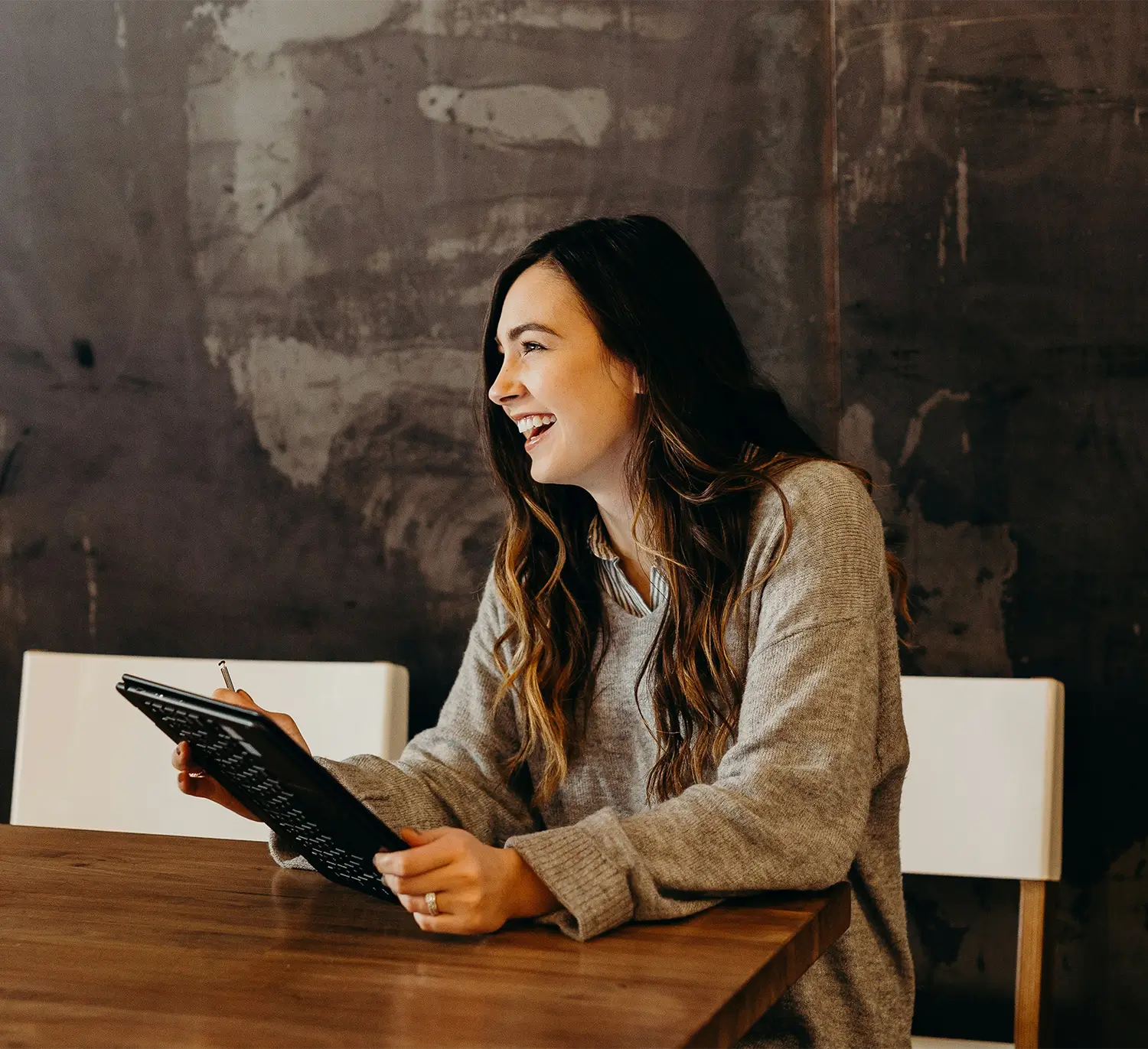 Woman sat in a co-working space with a tablet device, smiling at something out of shot.