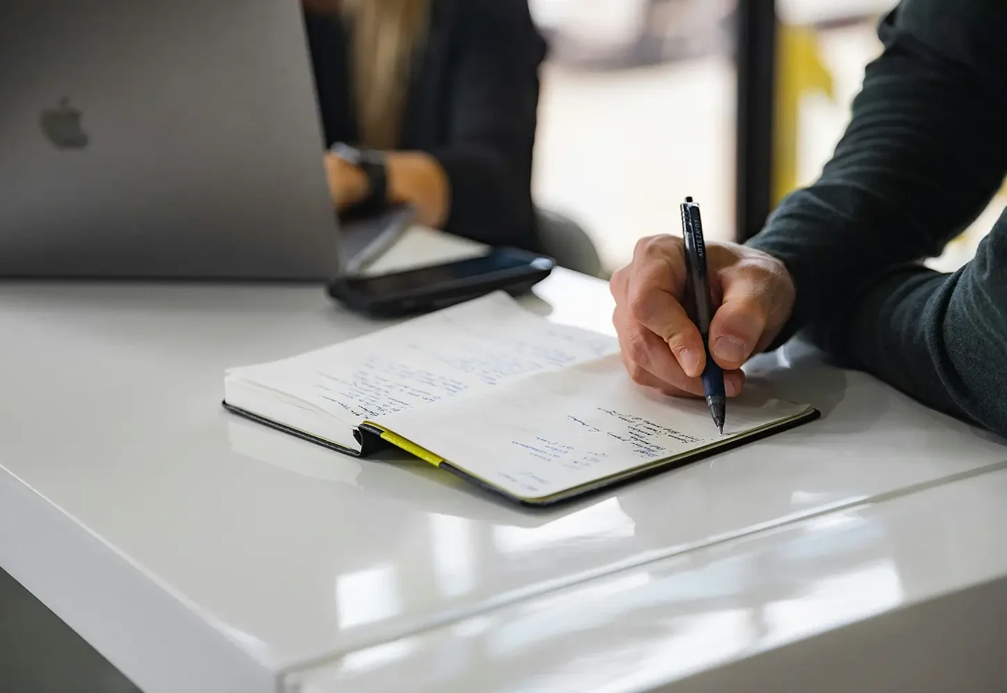 Close up of a hand holding a pen, writing in a notebook on a desk, with a person on a laptop in the background.